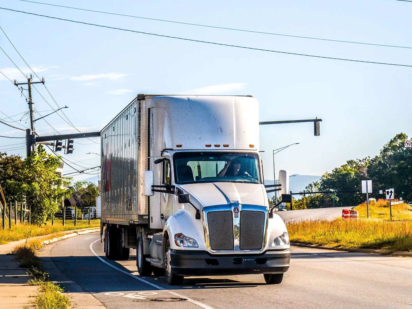 White semi-truck pulling a dry-van trailer around a curve at a signalized intersection, driver visible at the wheel, power lines overhead