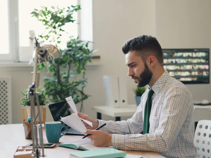 Man in a shirt and tie reading a printed document closely at a desk with a laptop and plants by a window