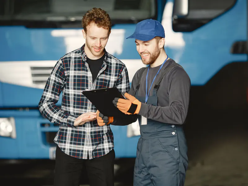 Mechanic in a cap going over a clipboard with a driver in front of a blue truck in a garage
