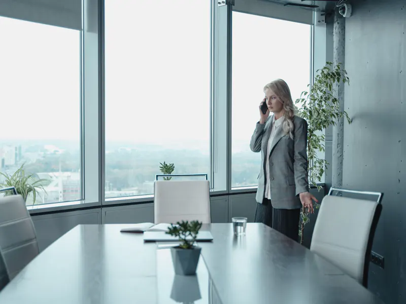 A woman in a gray blazer takes a phone call alone in the corner of an empty meeting room, a closed notebook on the table