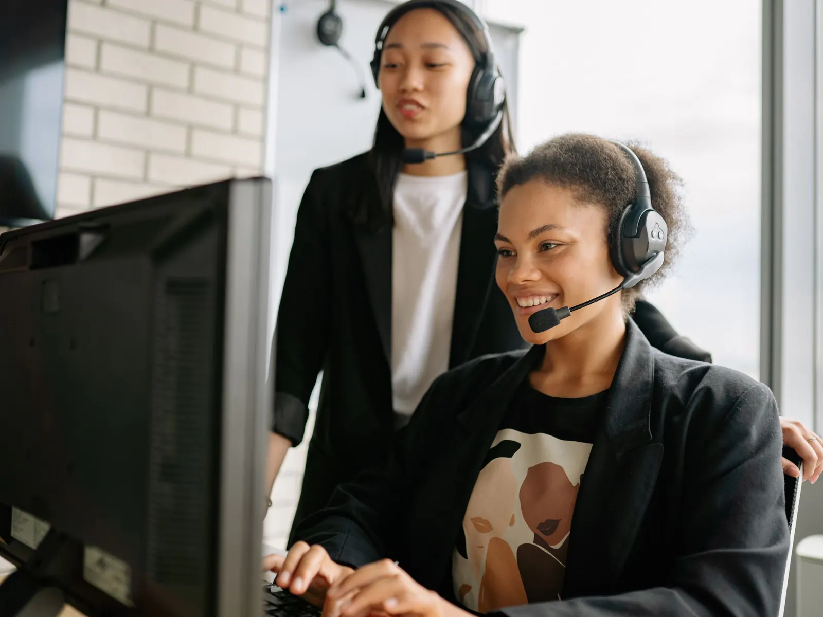 Smiling support agent wearing a headset at her computer while a colleague with a headset stands beside her