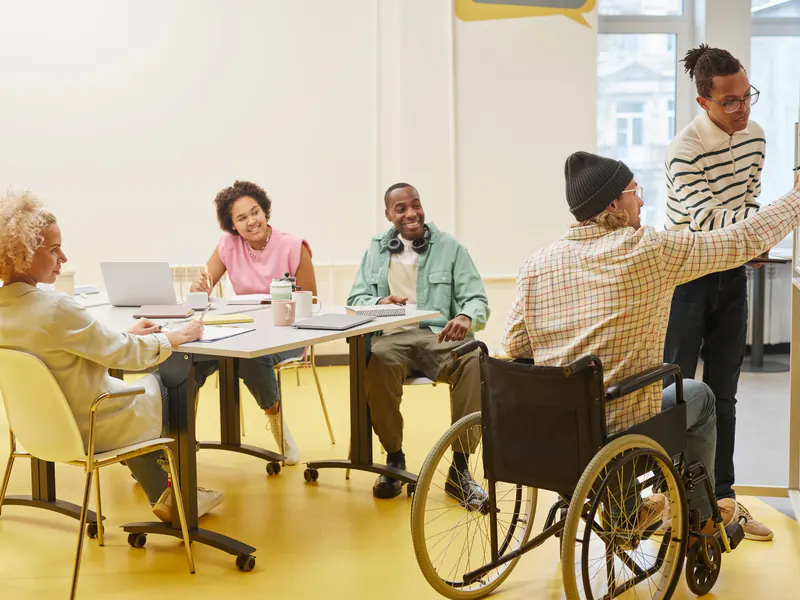 Diverse team of colleagues of different ages around a meeting table while a man in a wheelchair adds notes to a board