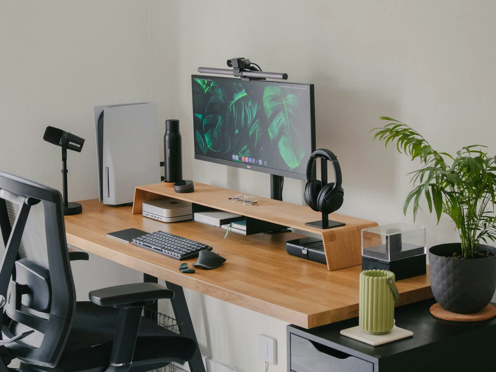 Tidy modern workspace with a monitor on a wooden riser, keyboard, microphone, headphones and a plant on a wood desk