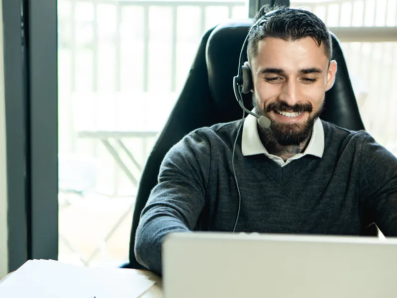 Smiling bearded man wearing a headset with a microphone working on a laptop beside a bright window