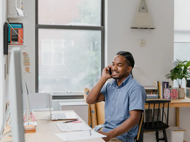 Smiling man in a blue short-sleeve shirt on a phone call at a desk with a laptop and papers