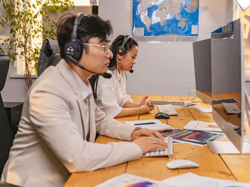 Two dispatchers wearing headsets working at computers on a shared desk, with a world map on the wall behind them