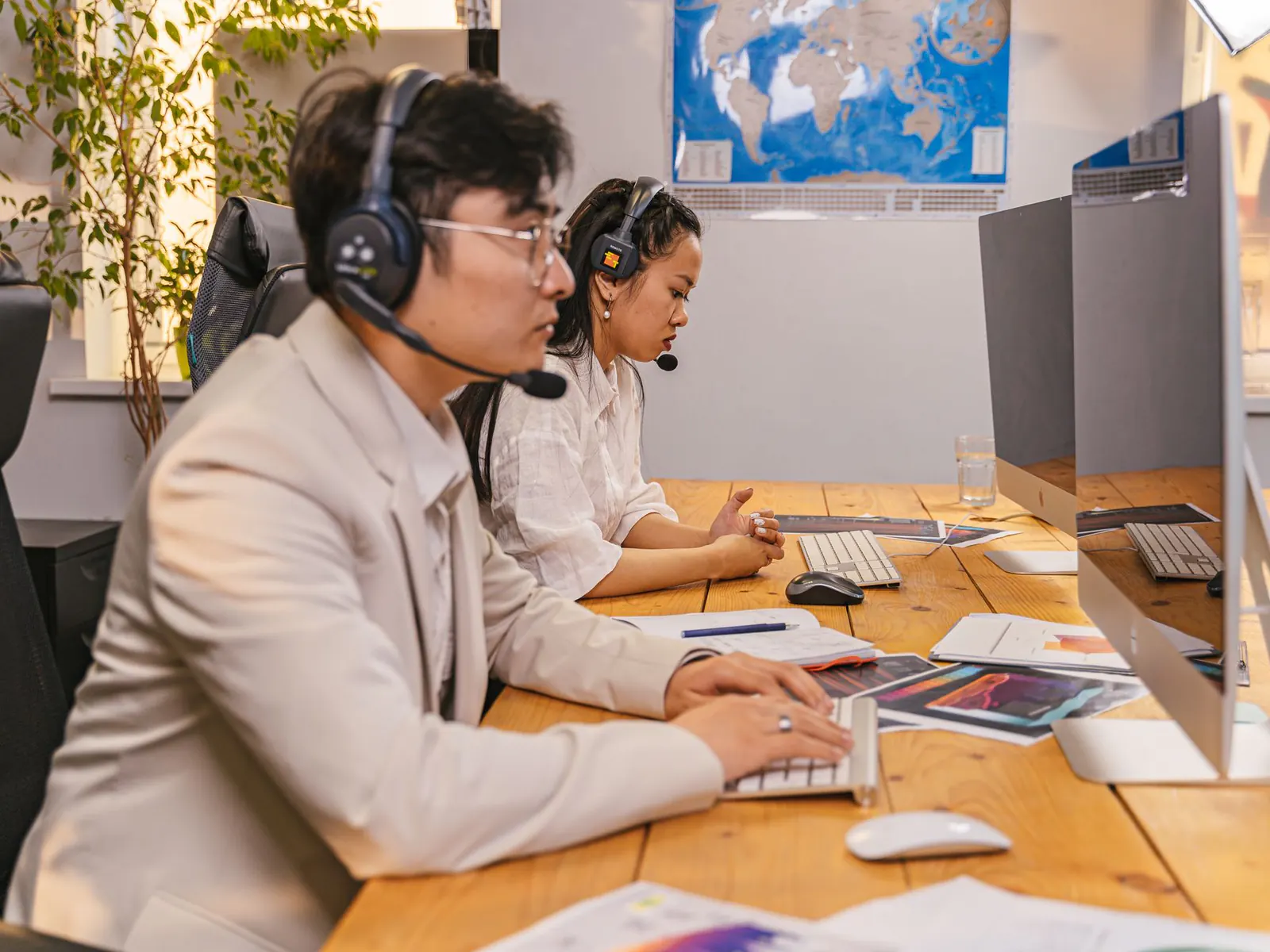 Two dispatchers wearing headsets working at computers on a shared desk, with a world map on the wall behind them