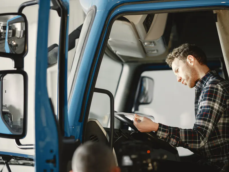 Man in a plaid shirt sitting in an open truck cab, reading paperwork at the steering wheel