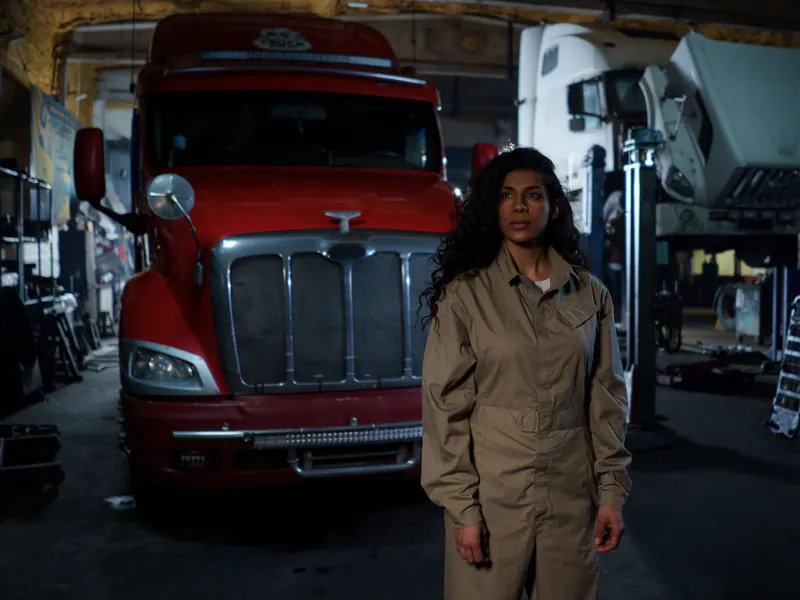 Woman in tan work coveralls standing beside the front of a red long-nose semi tractor inside a truck shop