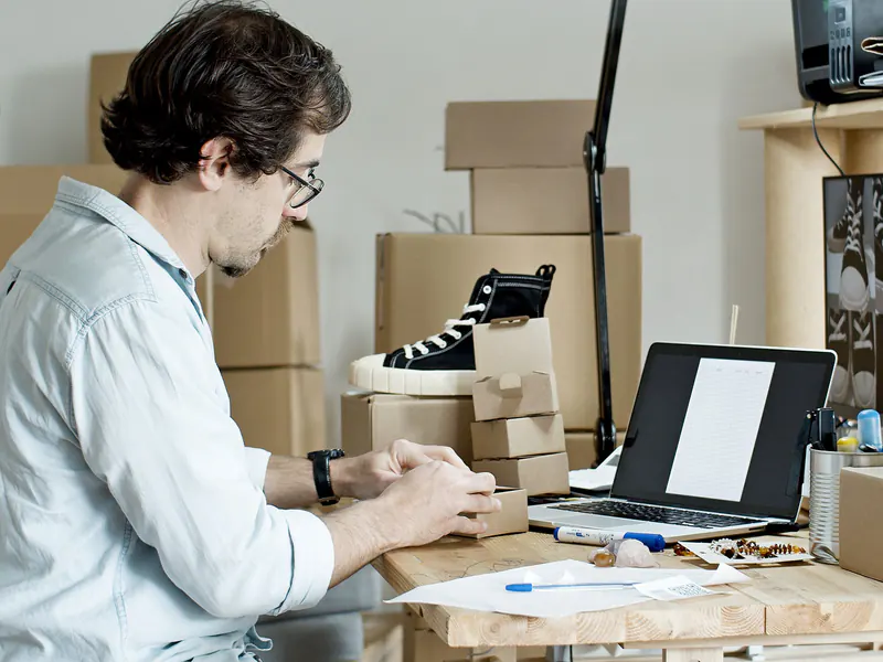 Online store owner at a desk with a laptop, preparing an order among sneakers and stacked shipping boxes