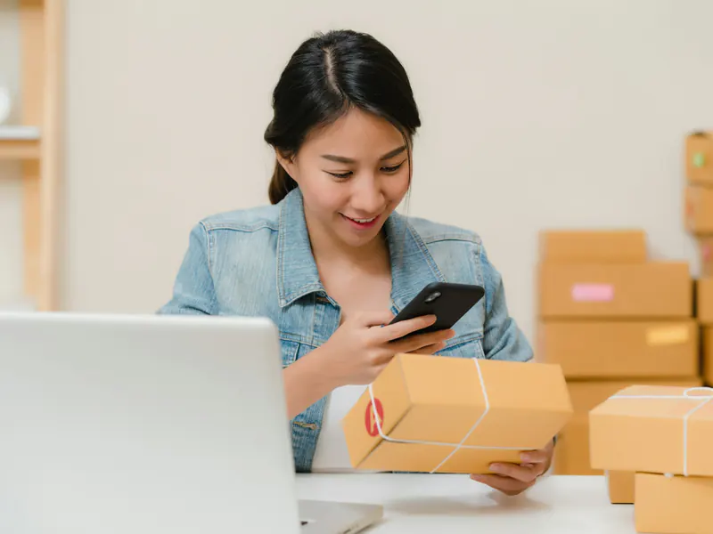 Smiling woman checking an order on her phone while holding a parcel, with a laptop and stacked cardboard boxes around her