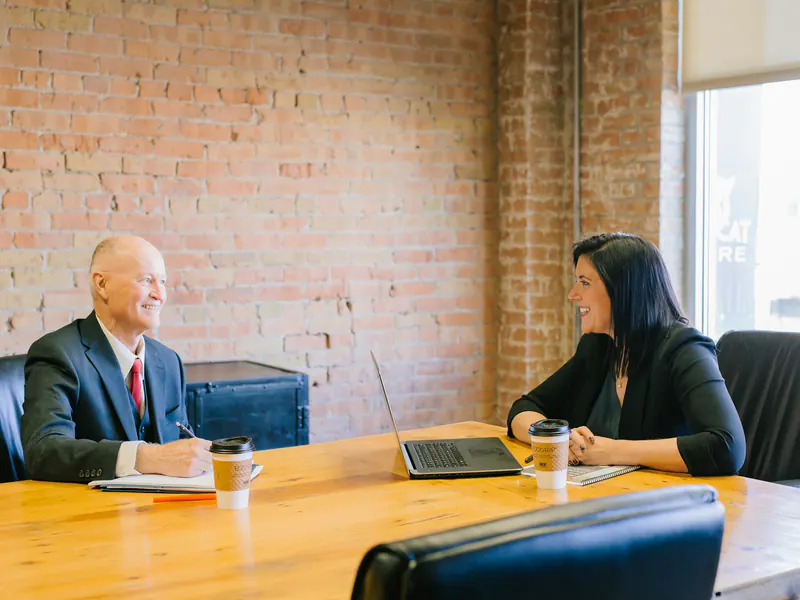 An older man in a suit and a woman in black talk across a wooden table with coffee in a brick-walled office