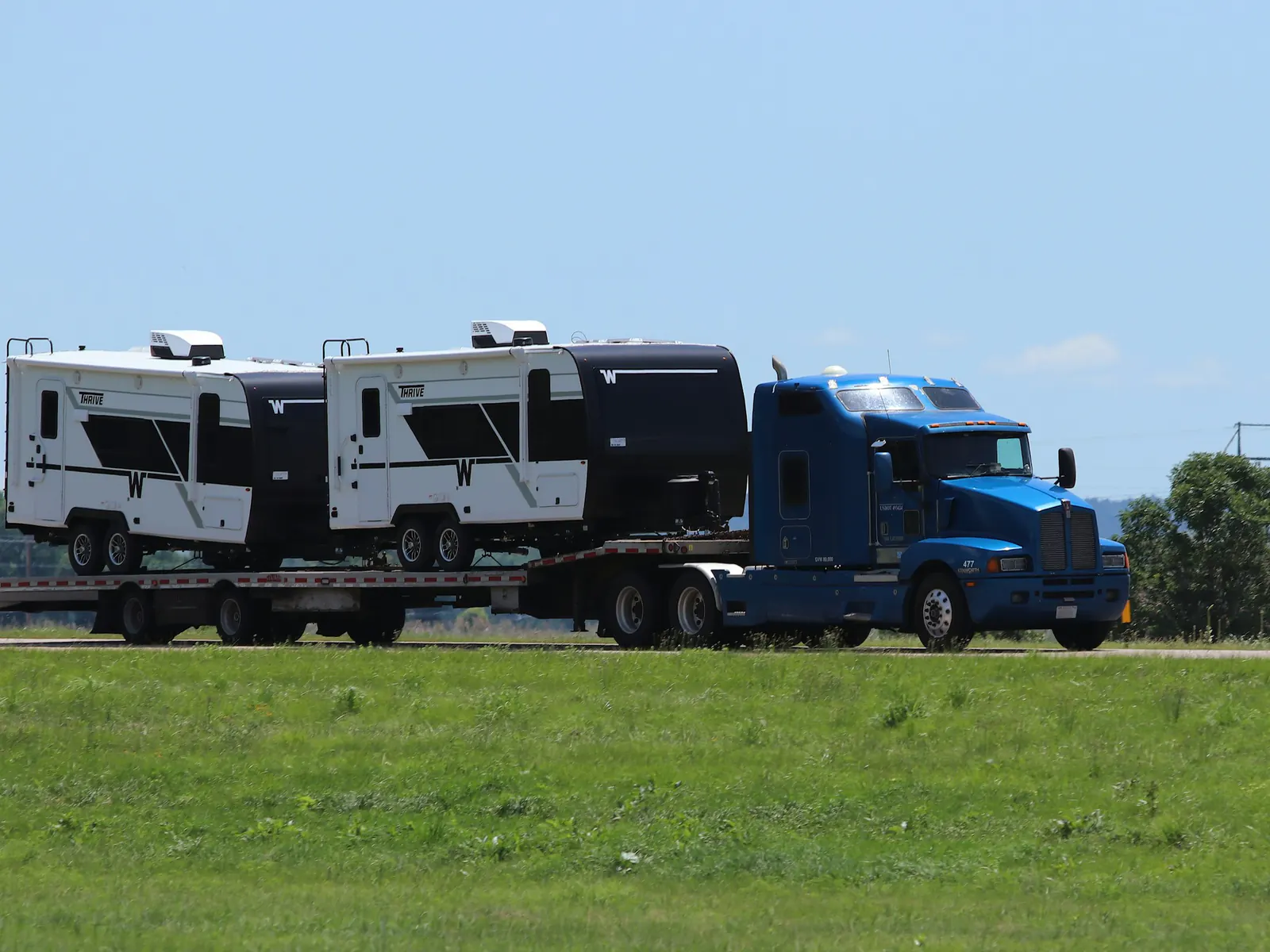 Blue semi truck hauling two travel trailers on a flatbed trailer past a green field