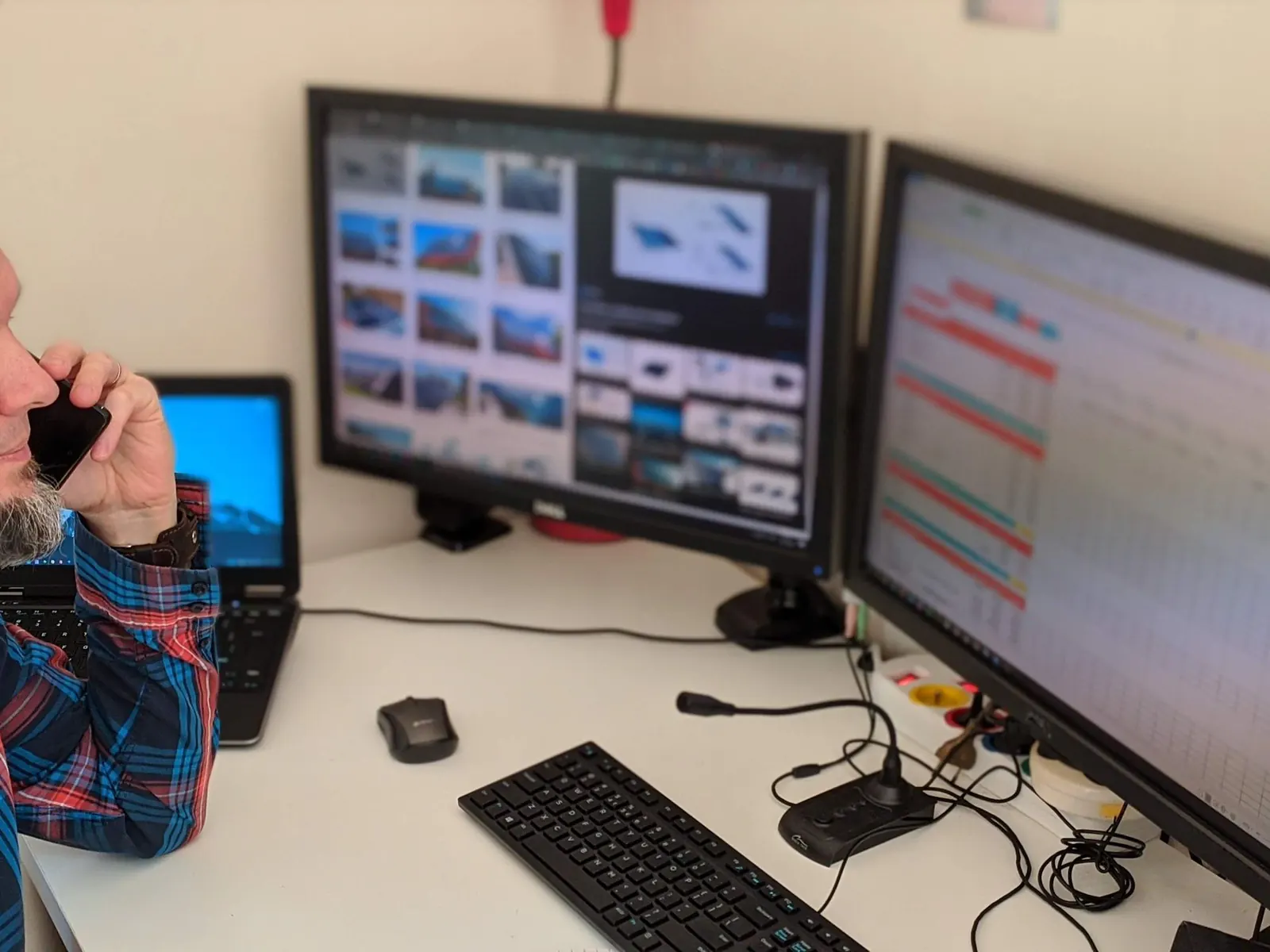 Man in a plaid shirt talking on a cell phone while watching two desktop monitors and a laptop at his office desk