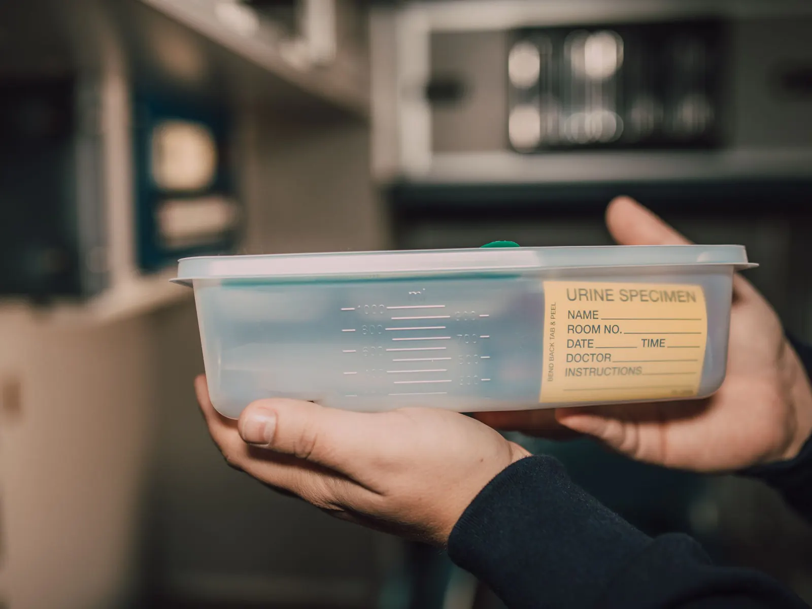 Two hands holding a clear plastic urine specimen collection container with a blank yellow label in a softly lit clinic room