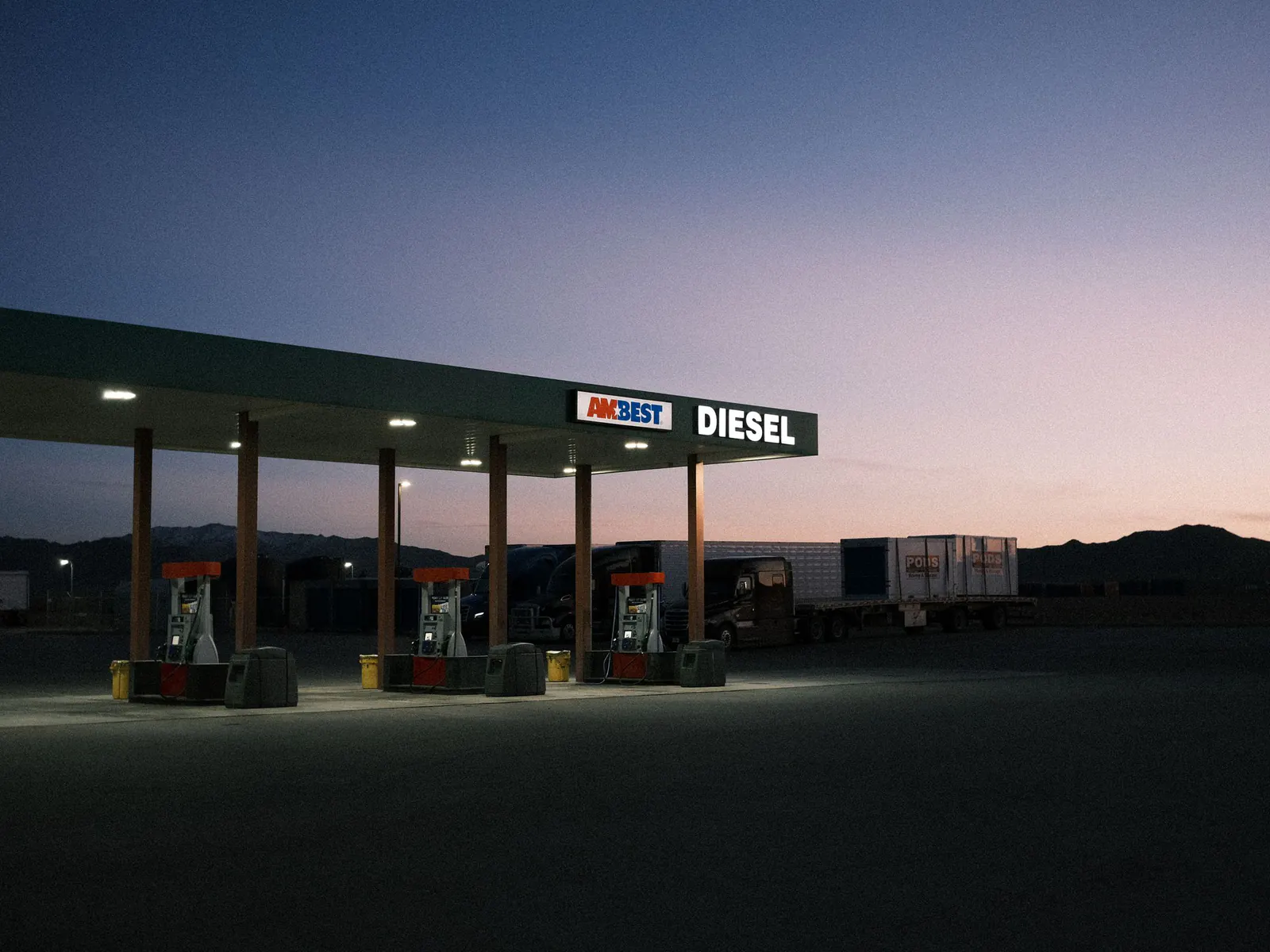 Truck stop diesel fuel island at dusk under a lit canopy, with parked semi trucks behind it and desert hills beyond