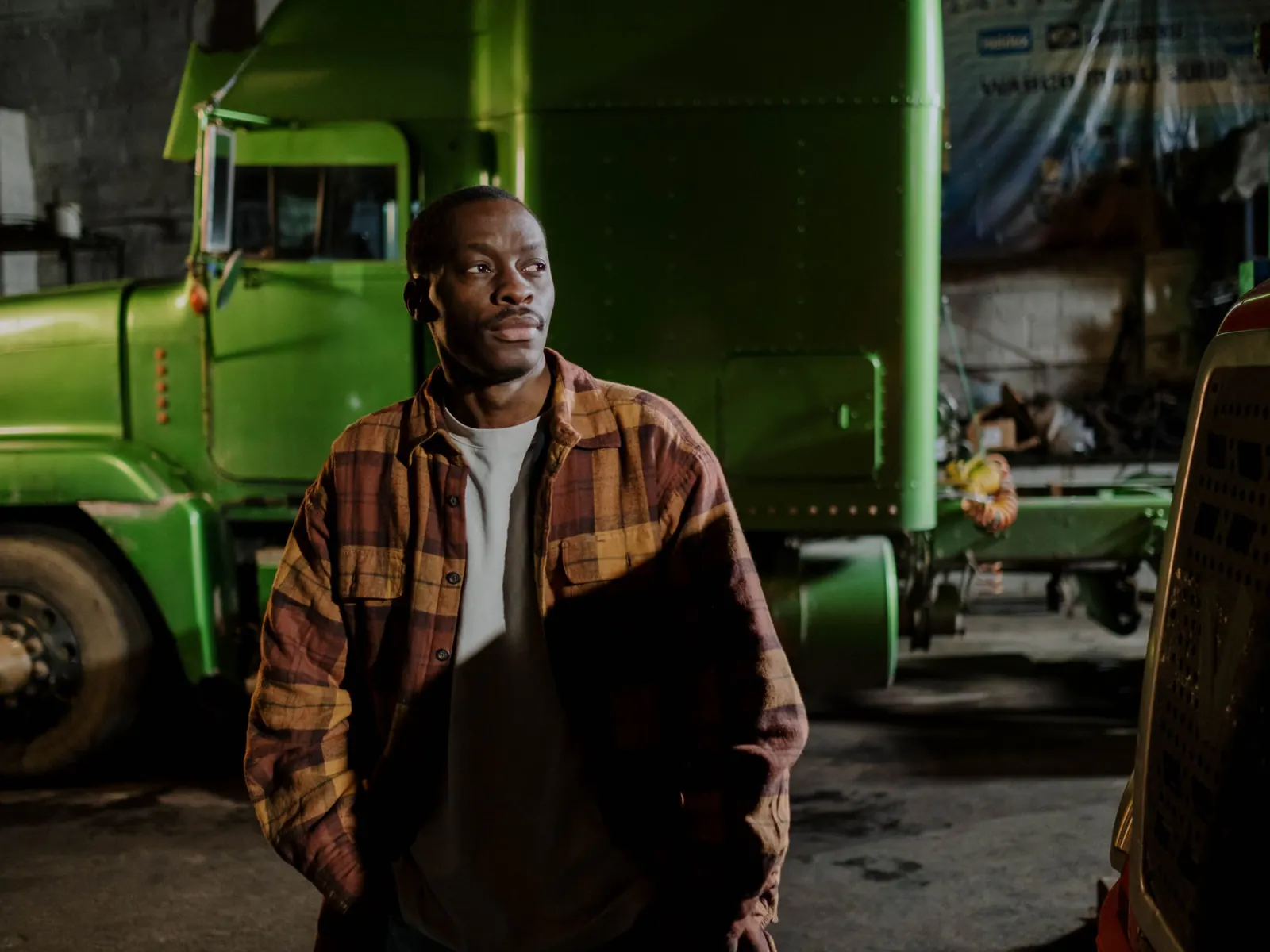 Man in a plaid flannel shirt standing beside the front of a green long-nose truck inside a dimly lit truck garage