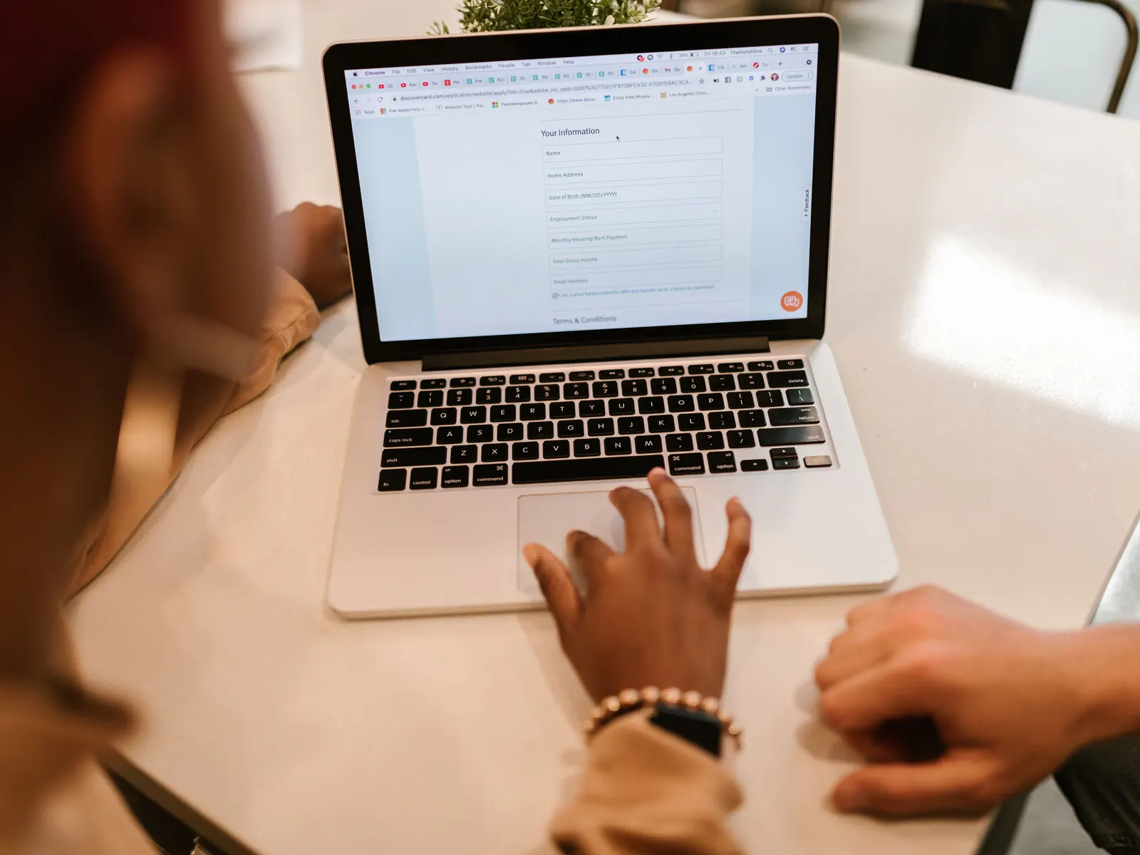 Hands on a laptop trackpad filling in an online form on screen, with a second person’s hand resting on the white table