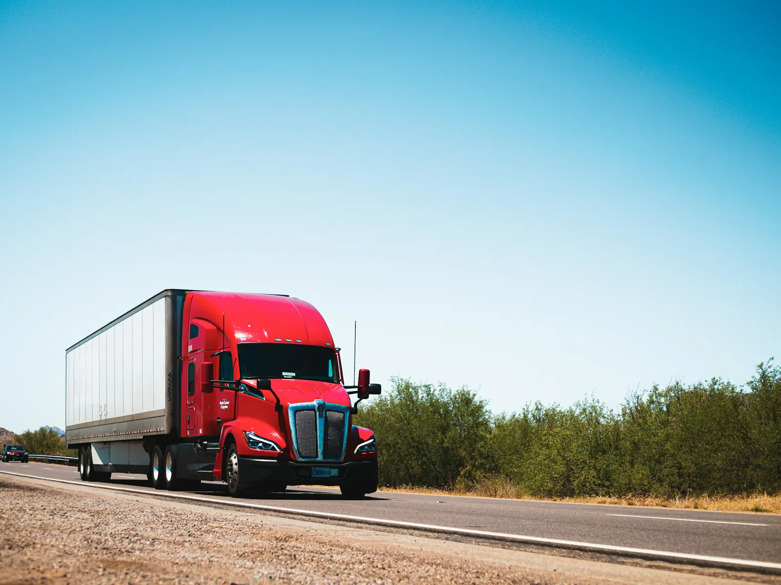 Late-model red long-nose semi tractor pulling a white dry van along a two-lane highway past desert scrub under a clear sky