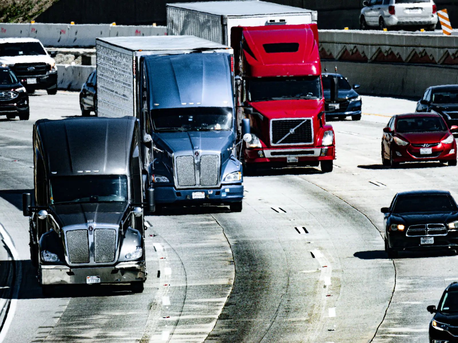 Three semi tractors pulling trailers in heavy traffic with cars on a curving multi-lane freeway on a sunny day