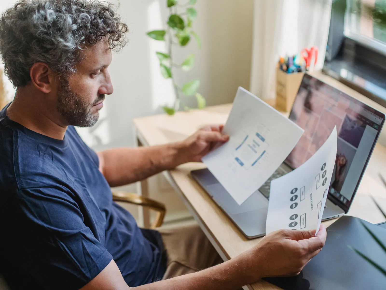 Middle-aged man with curly gray hair sitting at a home desk by a window, reviewing printed paperwork beside an open laptop