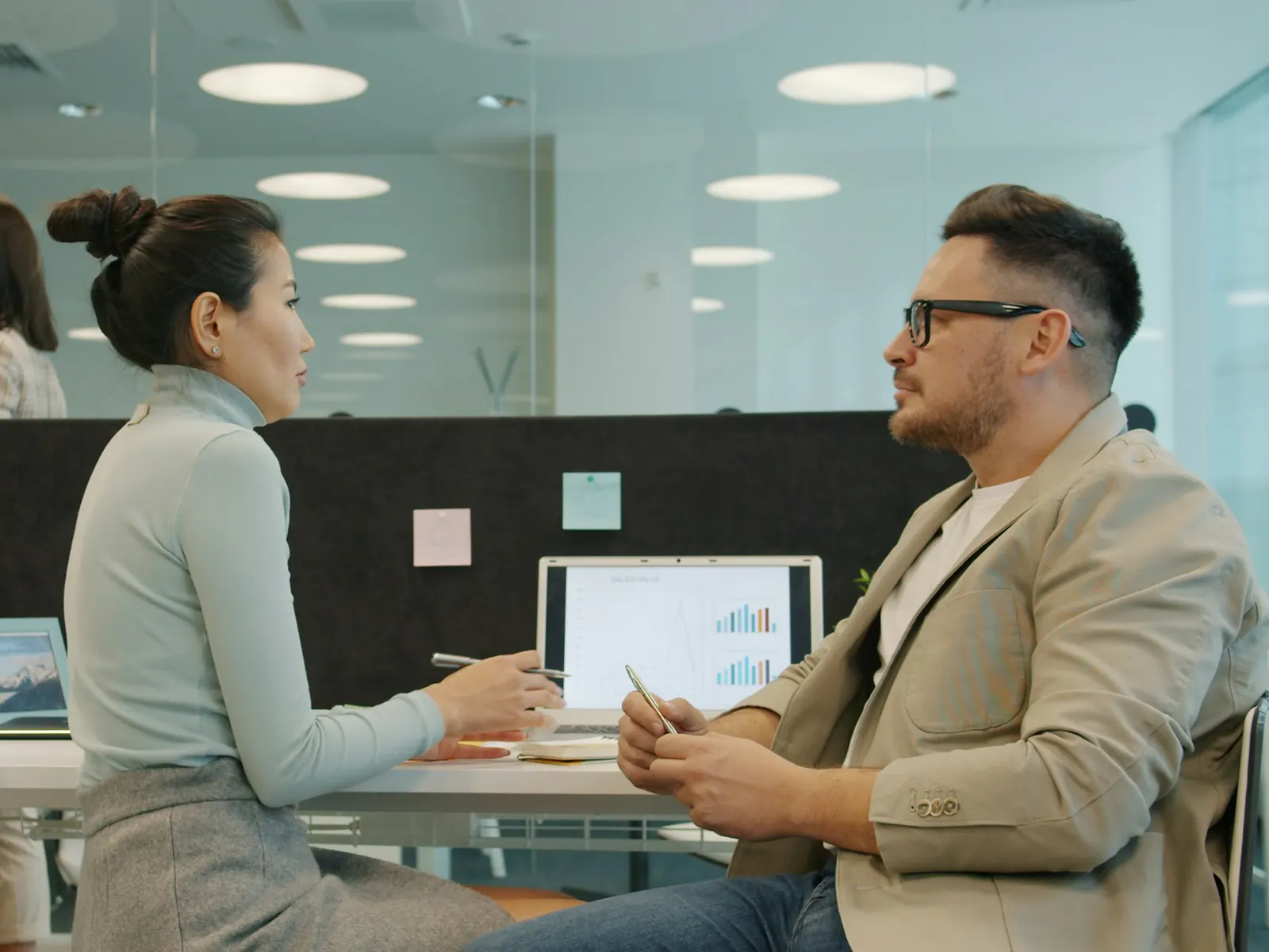 Two colleagues talking across a desk with a laptop showing charts in a bright, glass-walled open-plan office