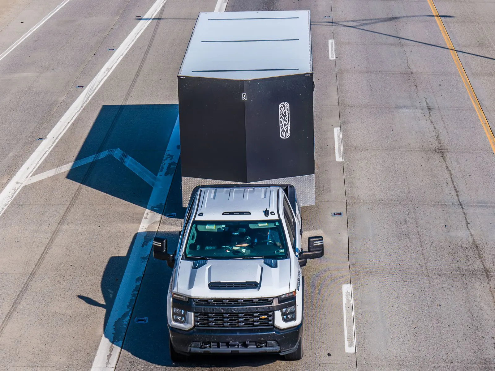 Aerial view of a silver heavy-duty pickup truck towing a black enclosed trailer down an empty highway
