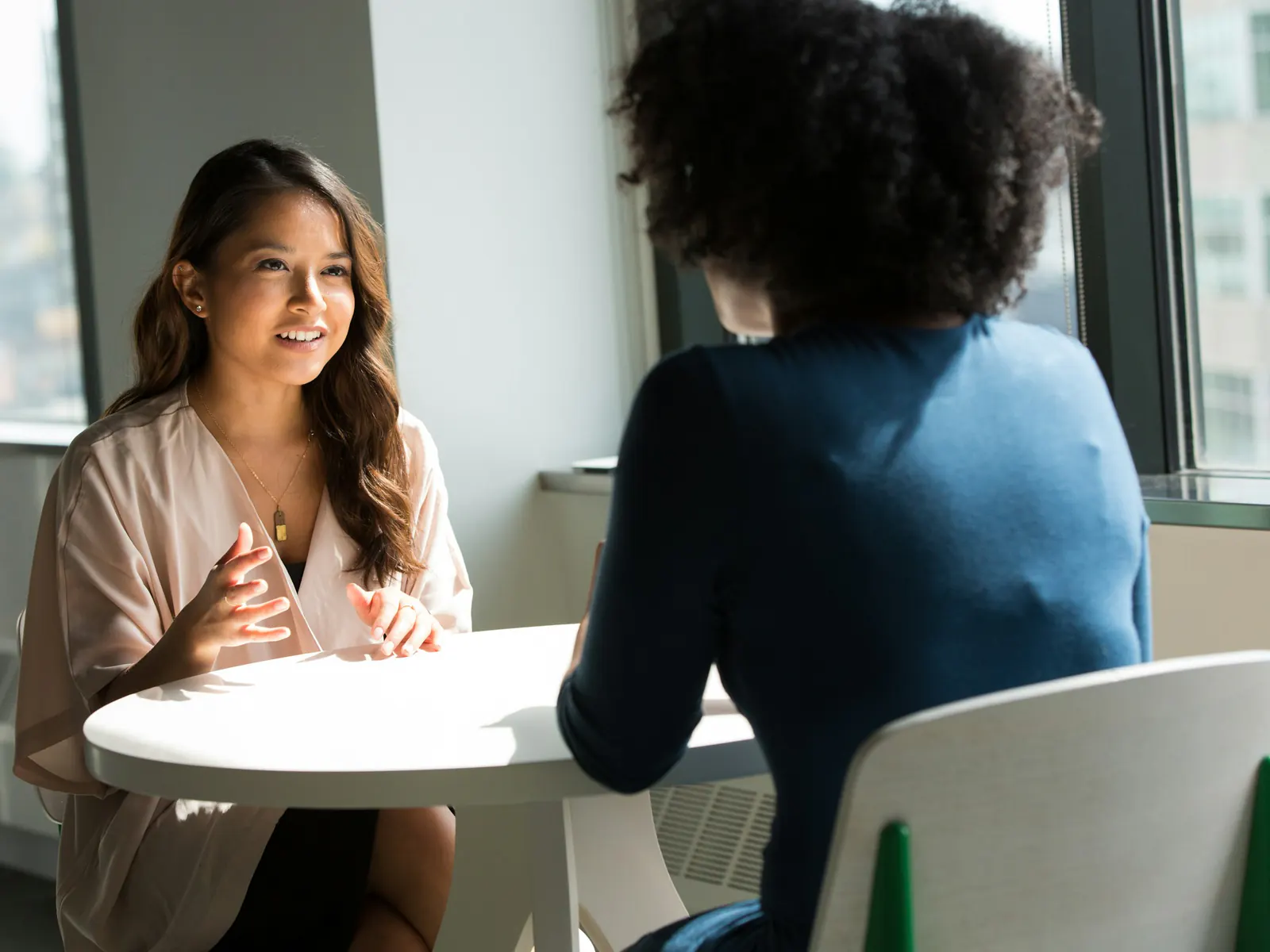 Two women talk across a small round table by a sunny office window; one smiles and gestures while the other listens.