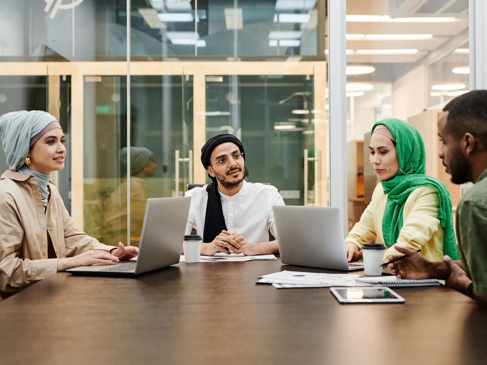 Four colleagues of different backgrounds, two women in headscarves, meeting at a conference table with laptops and coffee
