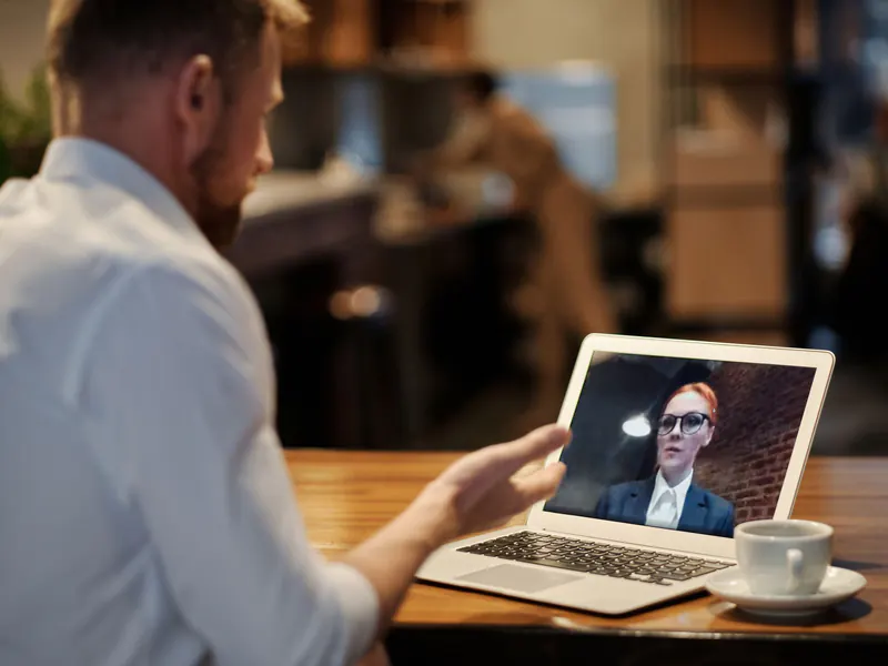 Man at a wooden table in warm evening light talking with a woman on his laptop screen, a cup of coffee beside it