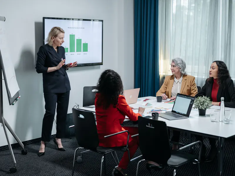 Woman presenting a bar chart on a wall screen to three seated women at a meeting table with laptops