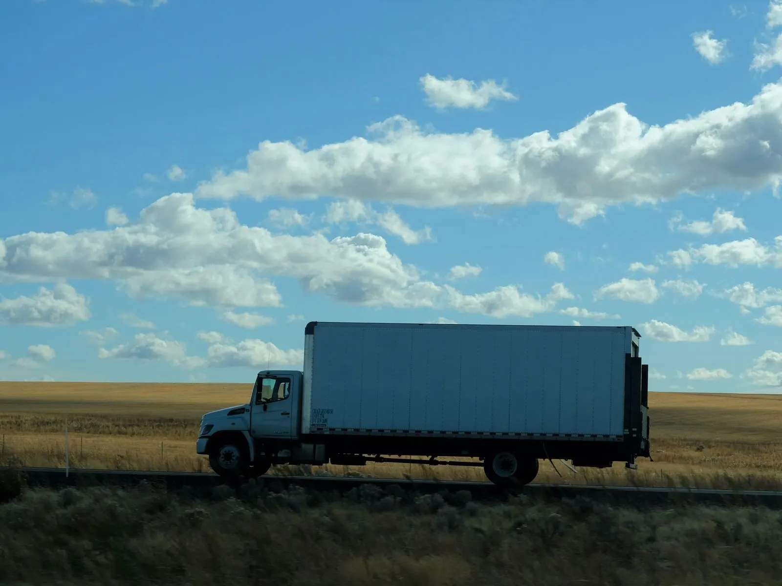 White straight box truck driving along a rural road past golden fields under a bright blue sky with rows of white clouds