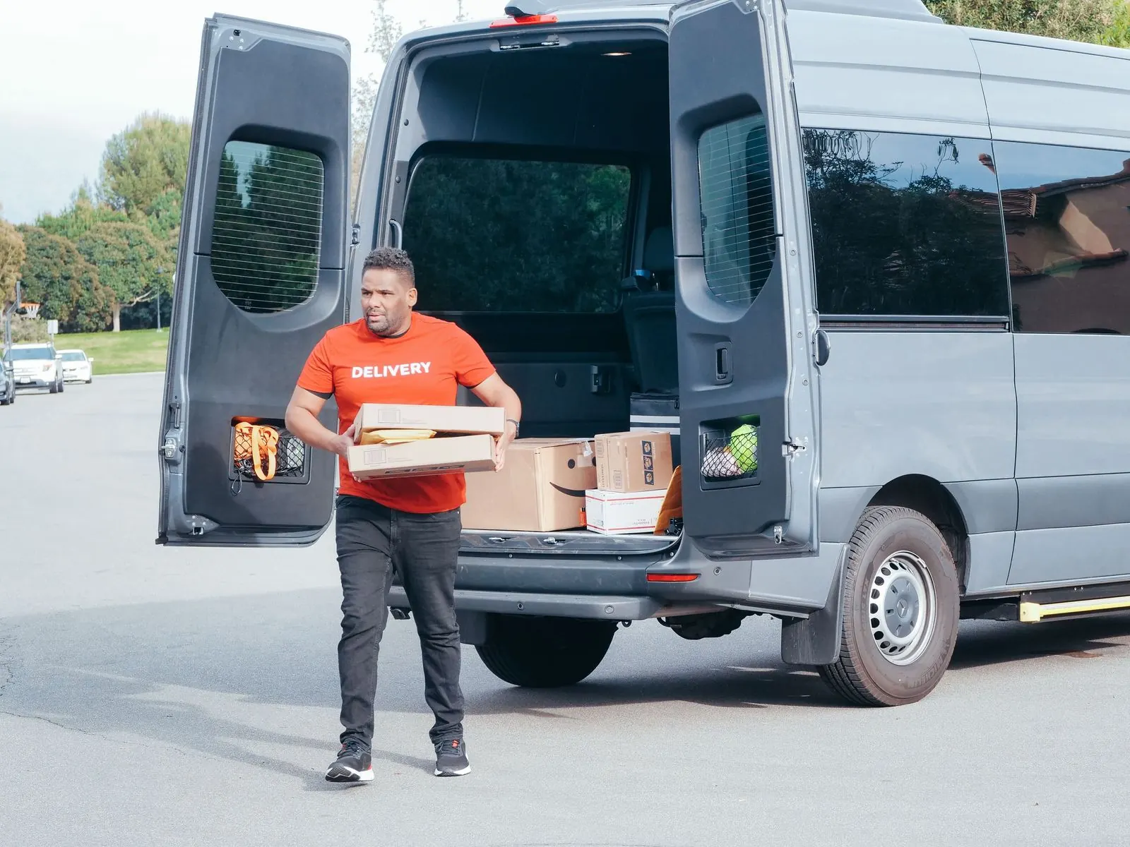 Delivery driver in an orange shirt carries boxes away from a gray high-roof cargo van with its rear doors open on a quiet street