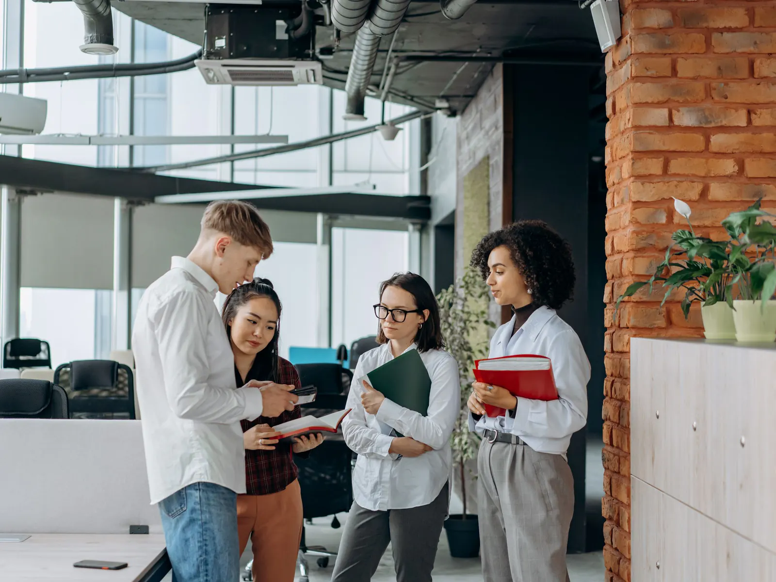 Four young colleagues stand in a loose huddle reading notebooks and folders in an open-plan loft office with brick walls and tall windows