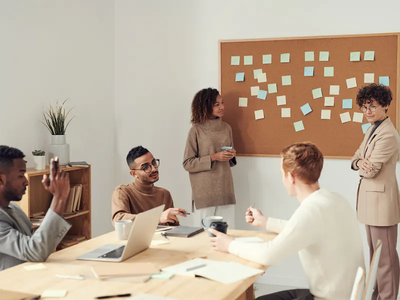 Five colleagues meet around a wooden table with a laptop and coffee while two women stand beside a corkboard covered in sticky notes