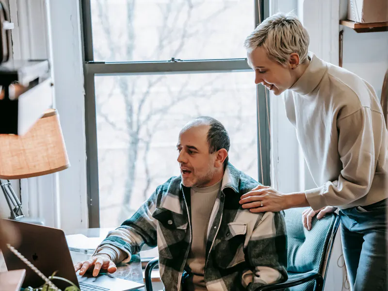 Smiling woman rests a hand on a seated colleague's shoulder as he works on a laptop at a desk by a window.
