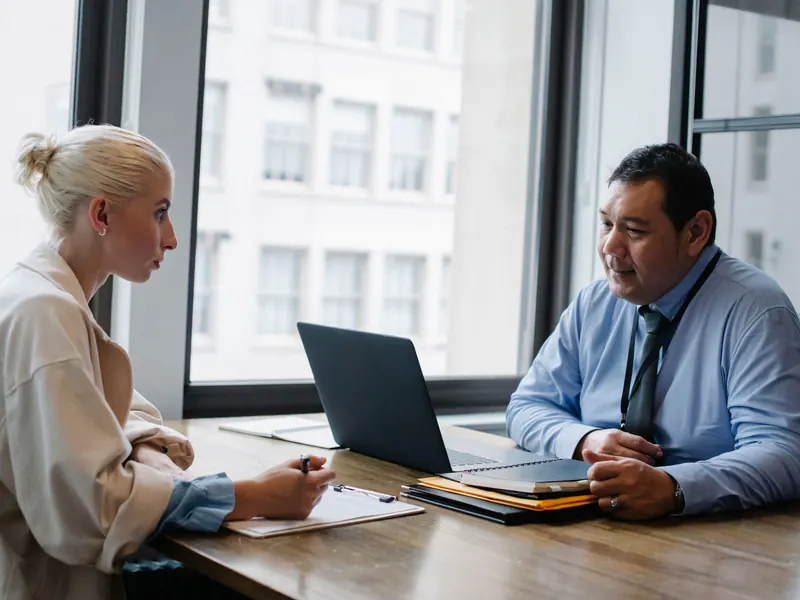 Man in a shirt and tie with a laptop interviews a blonde woman who takes notes across a wooden office table.