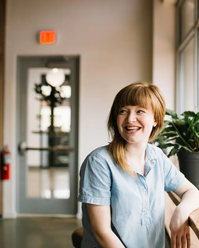 Smiling woman in a blue shirt sitting at a wooden desk in a sunlit office with a plant and an exit sign behind her