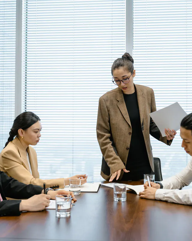 Woman in a tan blazer stands holding papers while advising three seated colleagues at a meeting room table