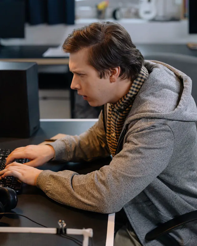 Young man in a grey hoodie typing at a computer desk in an office, headphones resting on the desk