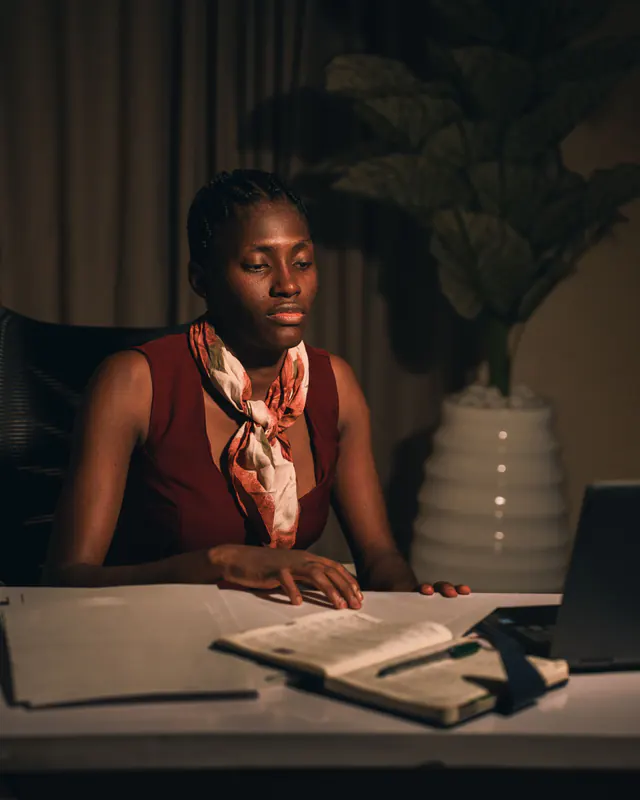 A woman in a red top and silk scarf sits at her desk in a dim office in the evening with a notebook