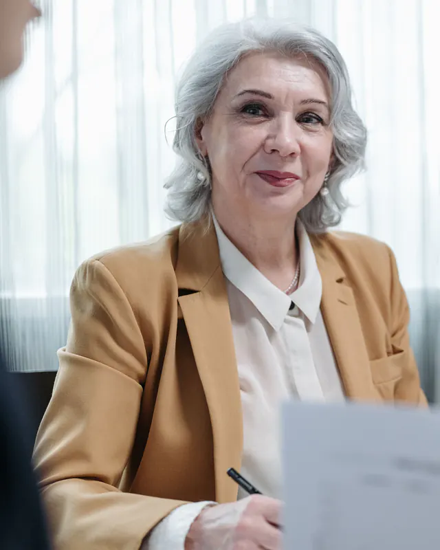 A woman with gray hair in a tan blazer sits at a meeting table with papers and a pen, smiling slightly