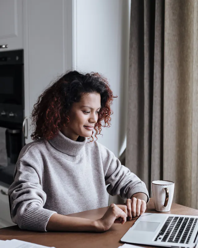 Woman in a gray turtleneck sweater working on a laptop at a table at home, with a mug and papers beside her