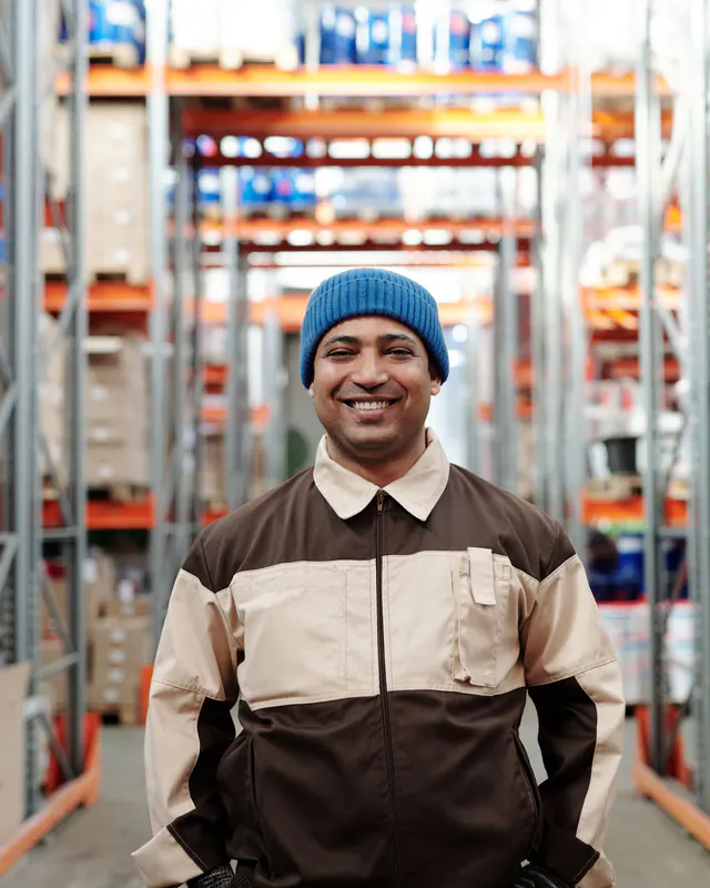 Smiling man in a blue beanie and two-tone work jacket stands in a warehouse aisle between tall pallet racks.