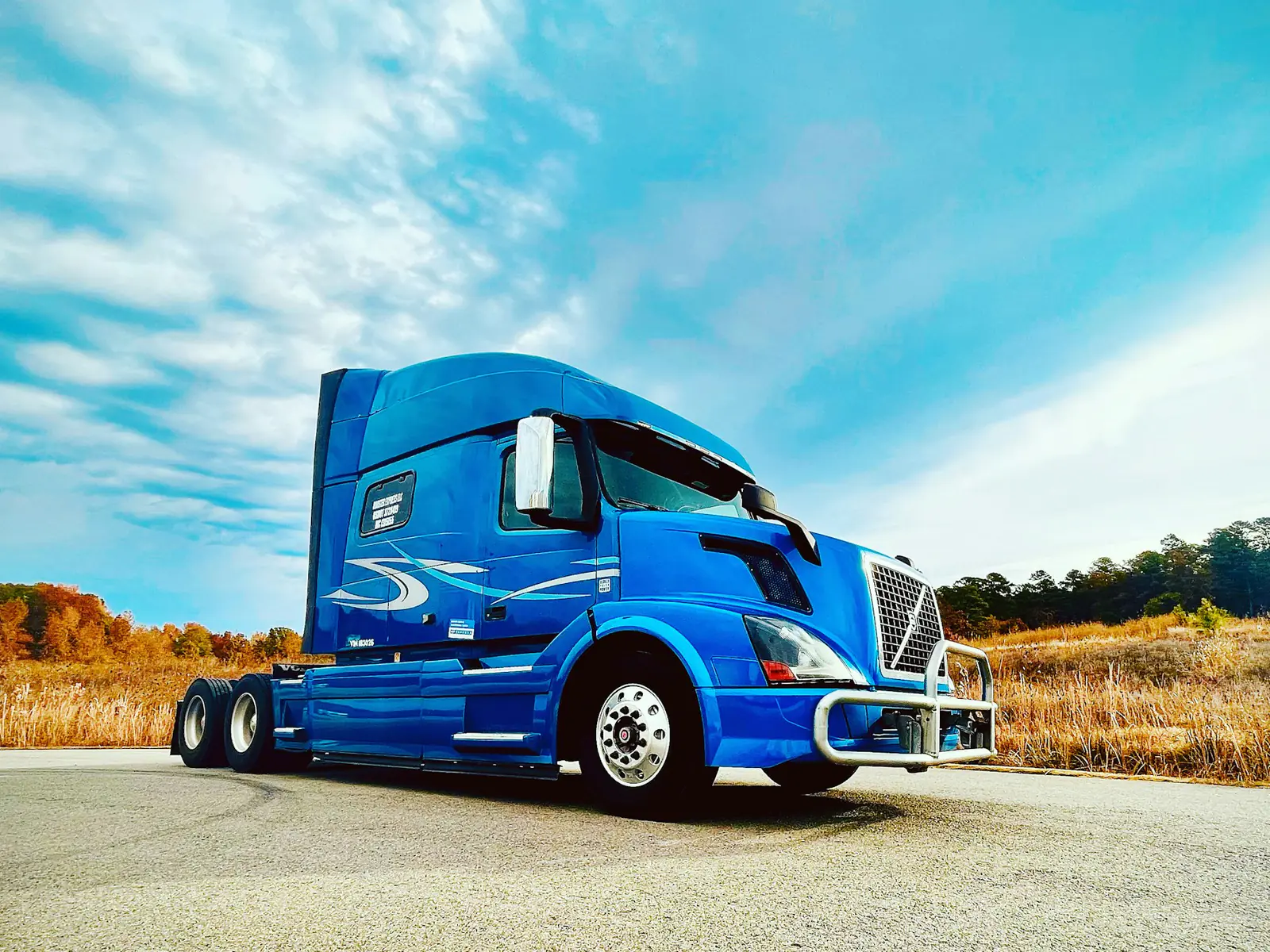 Blue semi tractor without a trailer parked on a road under a bright blue sky with autumn trees behind