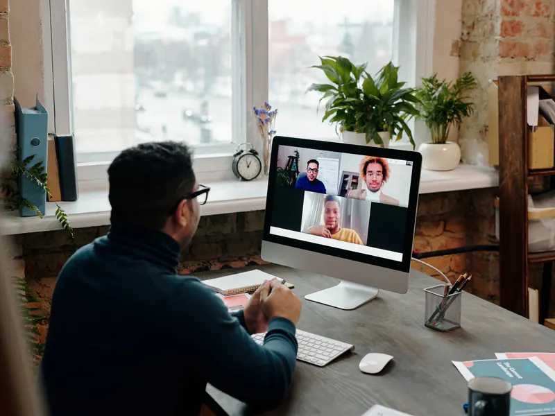 Man in glasses at a home office desk by a sunny window, on a video call with three colleagues shown on his desktop screen