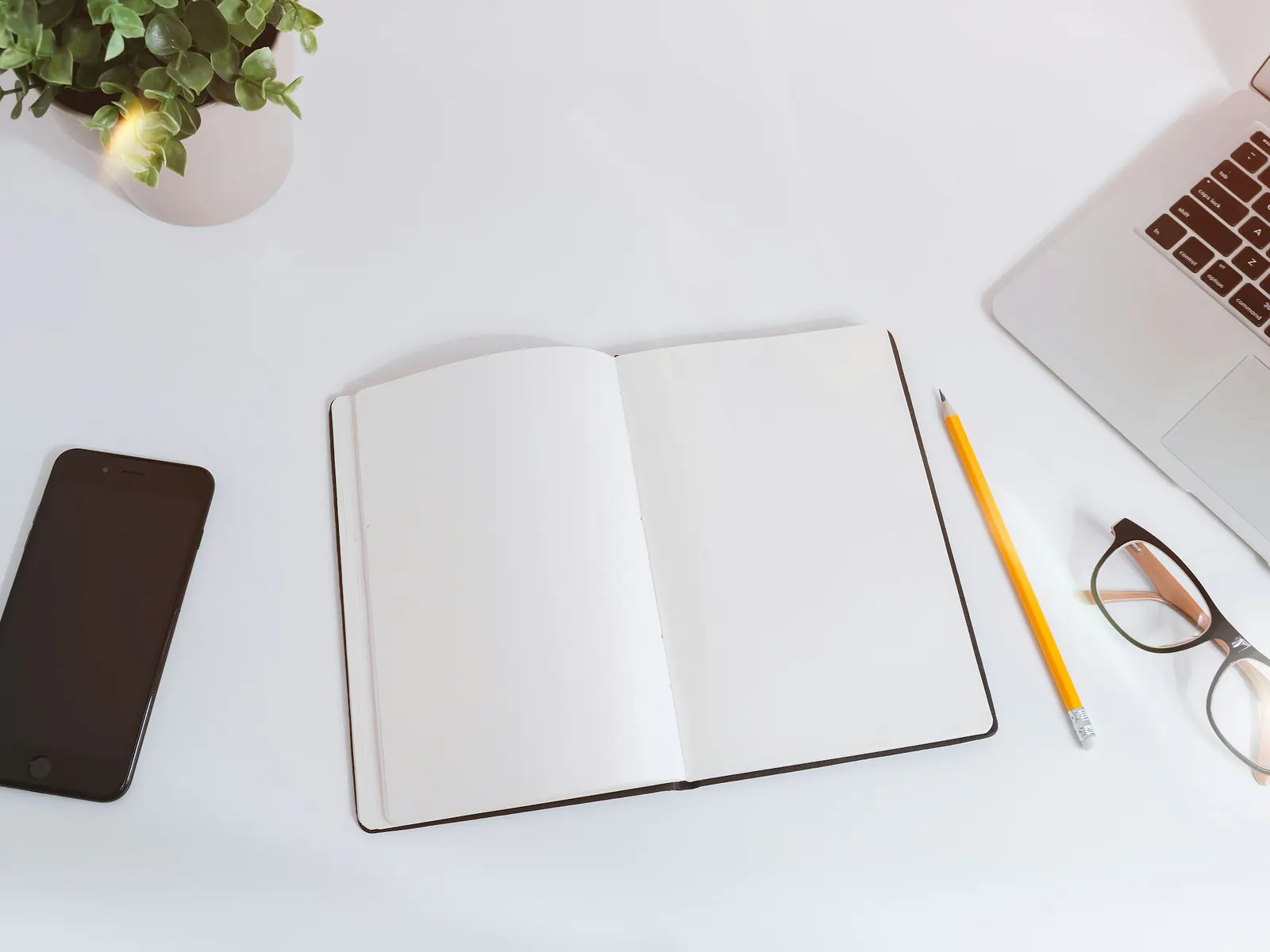 Open blank notebook with a pencil, glasses and a phone next to a laptop on a white desk, seen from above