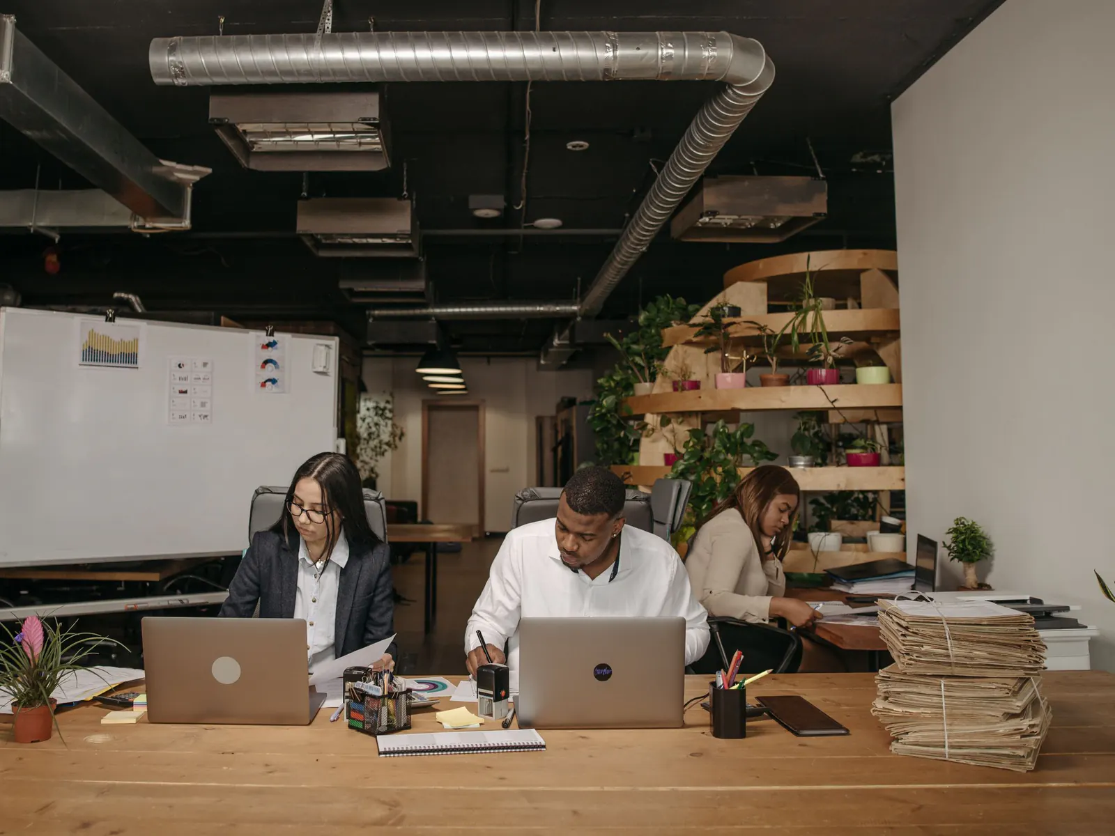 Three colleagues working at a long wooden desk with laptops, notebooks and stacks of paper files in an open office with a whiteboard behind