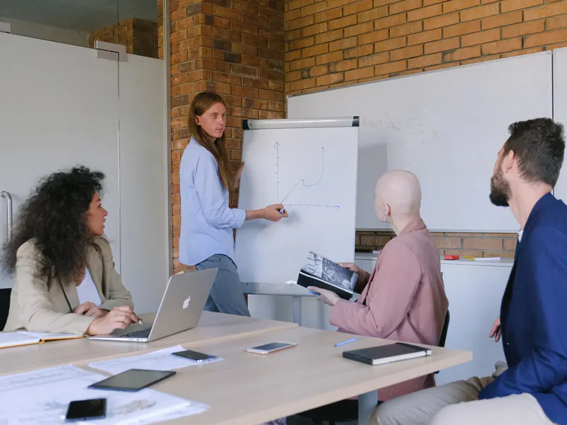 A woman sketches on a flipchart while three colleagues at a table with a laptop and notebooks watch, in a brick-walled meeting room
