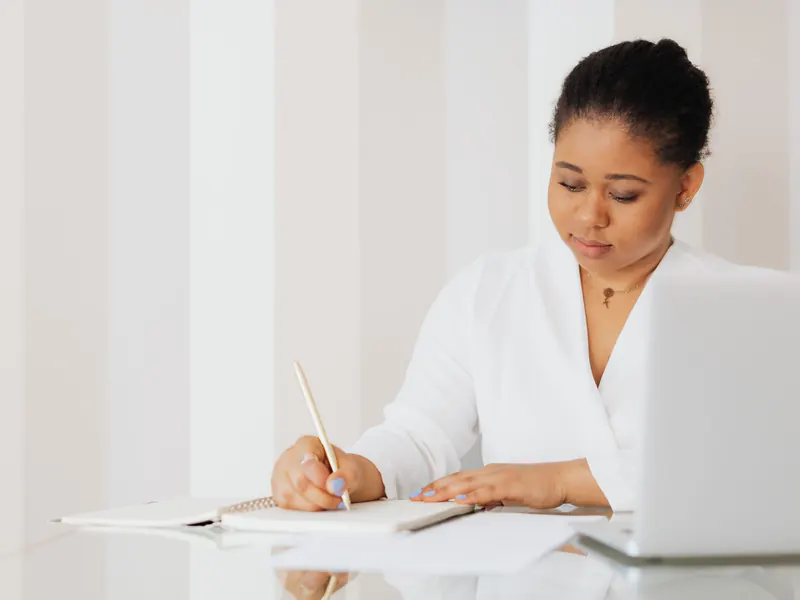 Woman in a white blouse writing notes in a notebook at a bright desk beside an open laptop